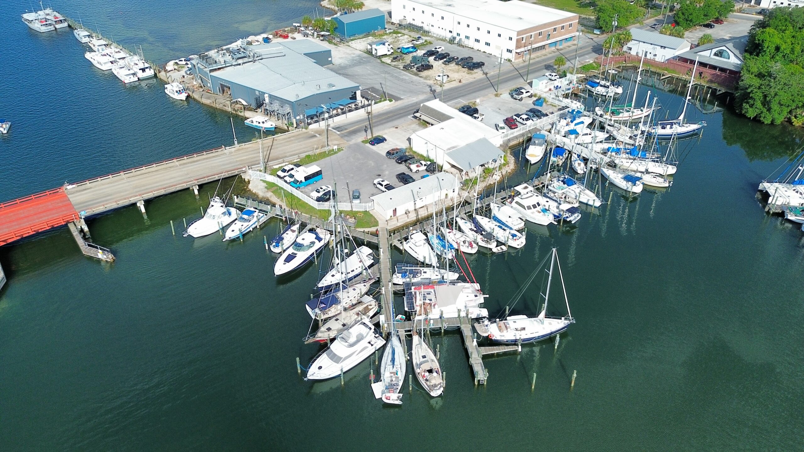 Aerial view of sailboats and powerboats docked at a marina beside a red bascule bridge, with waterfront commercial buildings along the road.
