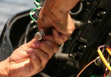 Hands working on the wiring of an outboard motor with a small cylindrical connector, with water visible in the background.