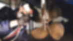 Close-up, blurry view of bronze boat propellers on a shaft, with a person partly visible in the background.