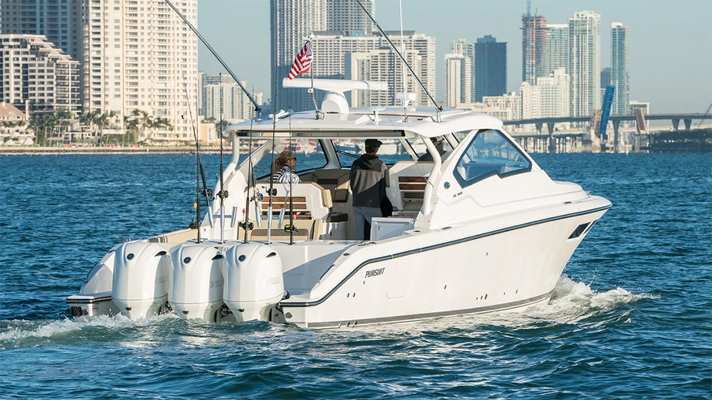 White Pursuit sport cruiser with triple outboard motors and rod holders cruising near a high-rise waterfront city, likely Miami.