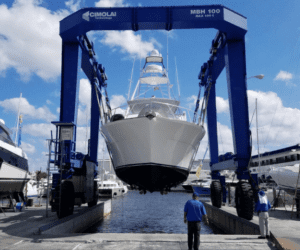 White sportfish boat being lifted by a blue Cromelin travel lift at a boatyard, with a worker on the dock below.