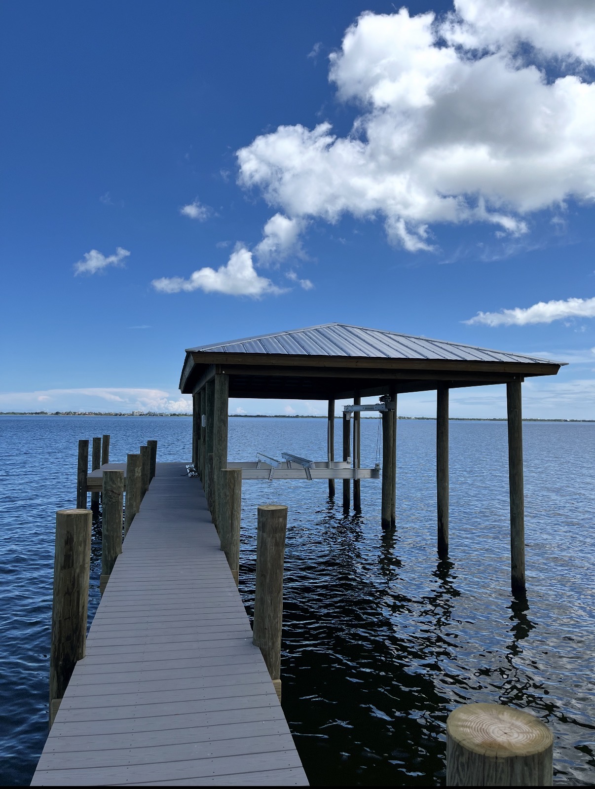 Covered boat dock with a metal-roofed boat lift and composite decking extending over the water, with a distant shoreline under a blue sky.