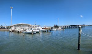 Docked white boats and a waterfront building at a marina with a long pier stretching across the calm water.
