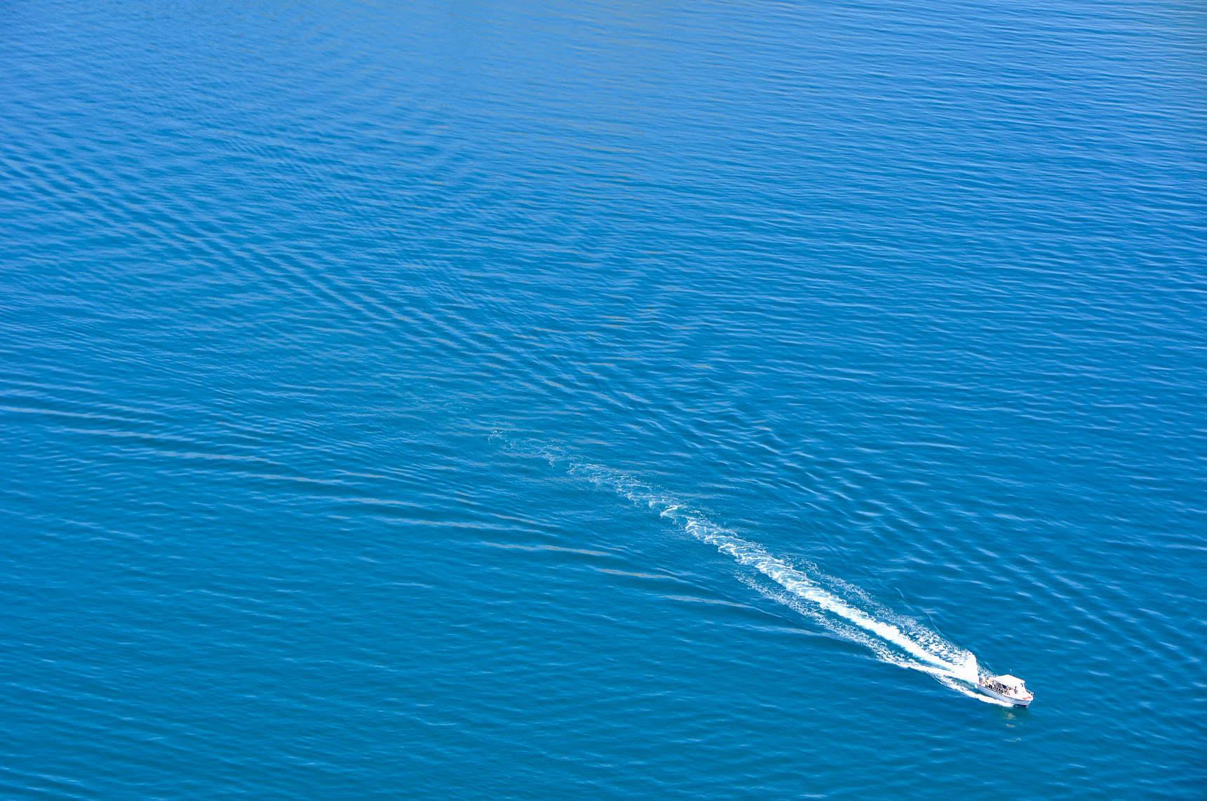 Small white passenger boat with a trailing wake crossing calm blue water, photographed from above.