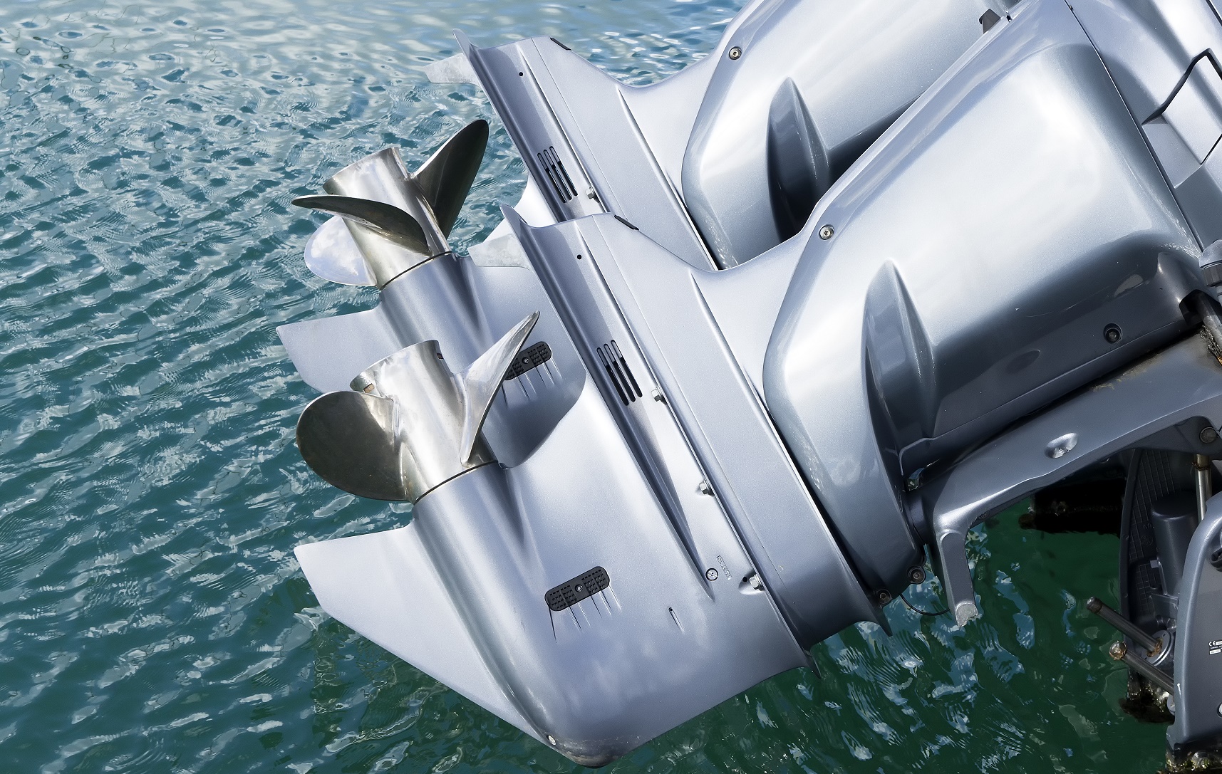 Twin silver outboard motors with stainless steel propellers mounted on a boat above green water.