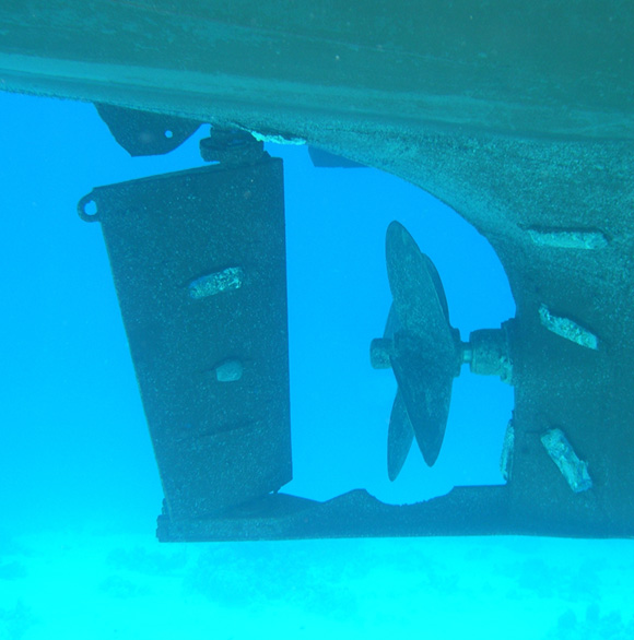 Underwater view of a boat's propeller and rudder beside the hull in clear blue water.