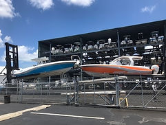 Two powerboats displayed in a dry-stack storage yard with racks of boats behind a chain-link fence.