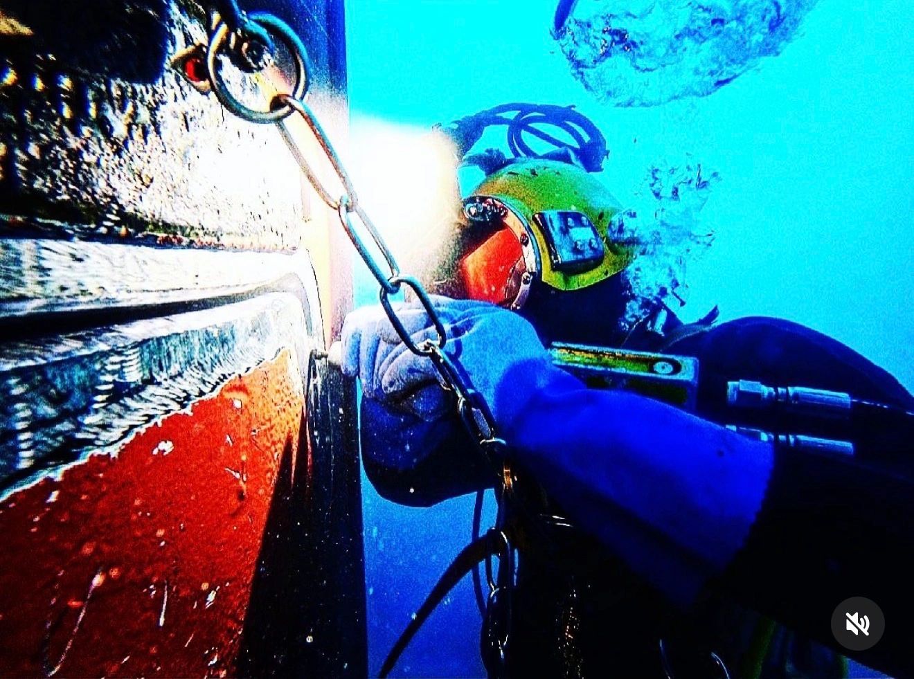 Diver in a green helmet working on a red-hulled boat's underwater hull with a chain and ring hanging alongside.