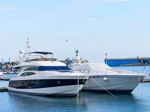 Two white motor yachts moored side by side in a harbor with a breakwater behind them.