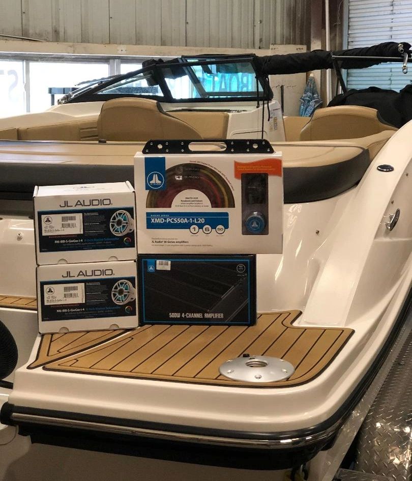 JL Audio marine subwoofers, a speaker kit, and a 4-channel amplifier set on the deck of a boat in a showroom.