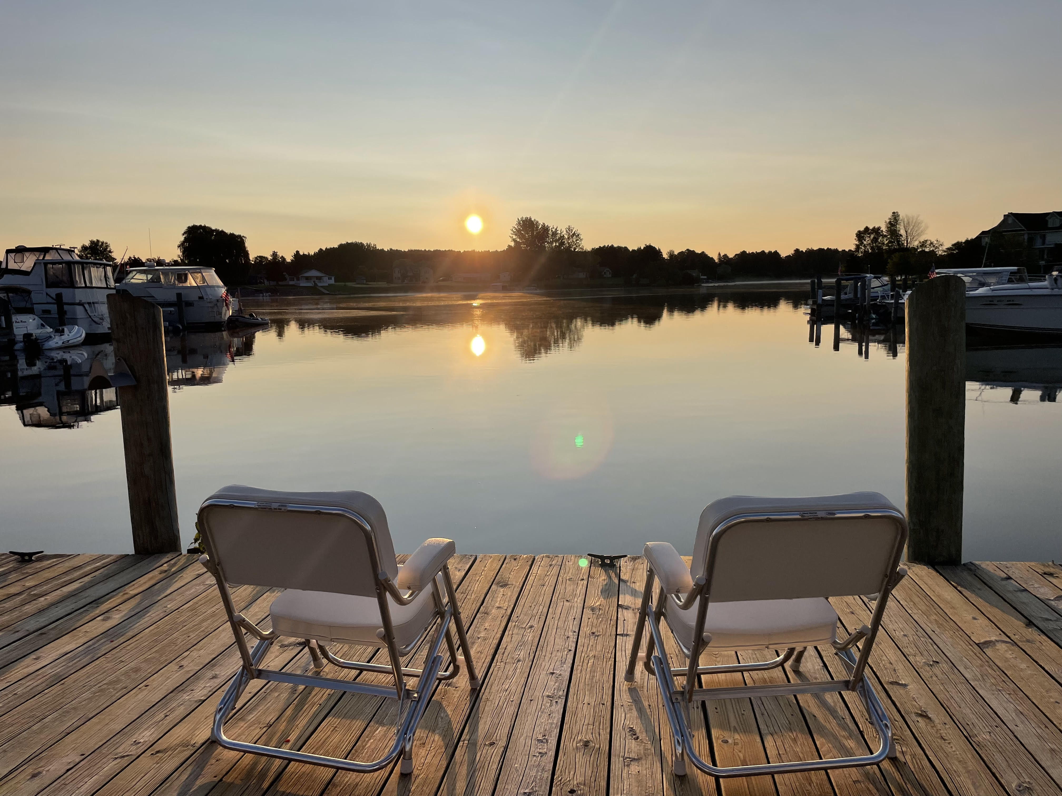 Two white folding deck chairs on a wooden dock facing a calm waterway with moored cabin cruisers at sunrise.