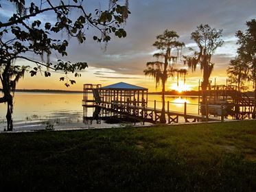 Covered wooden boat dock with a boat lift at sunset on a calm lake lined with cypress trees.