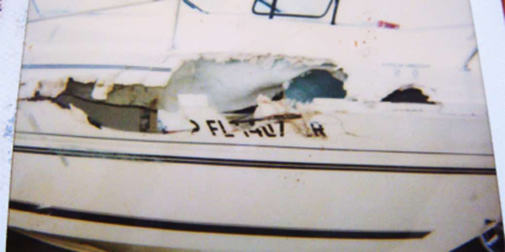 Damaged white sailboat hull with a large gaping hole in the fiberglass and partially legible Florida registration lettering.