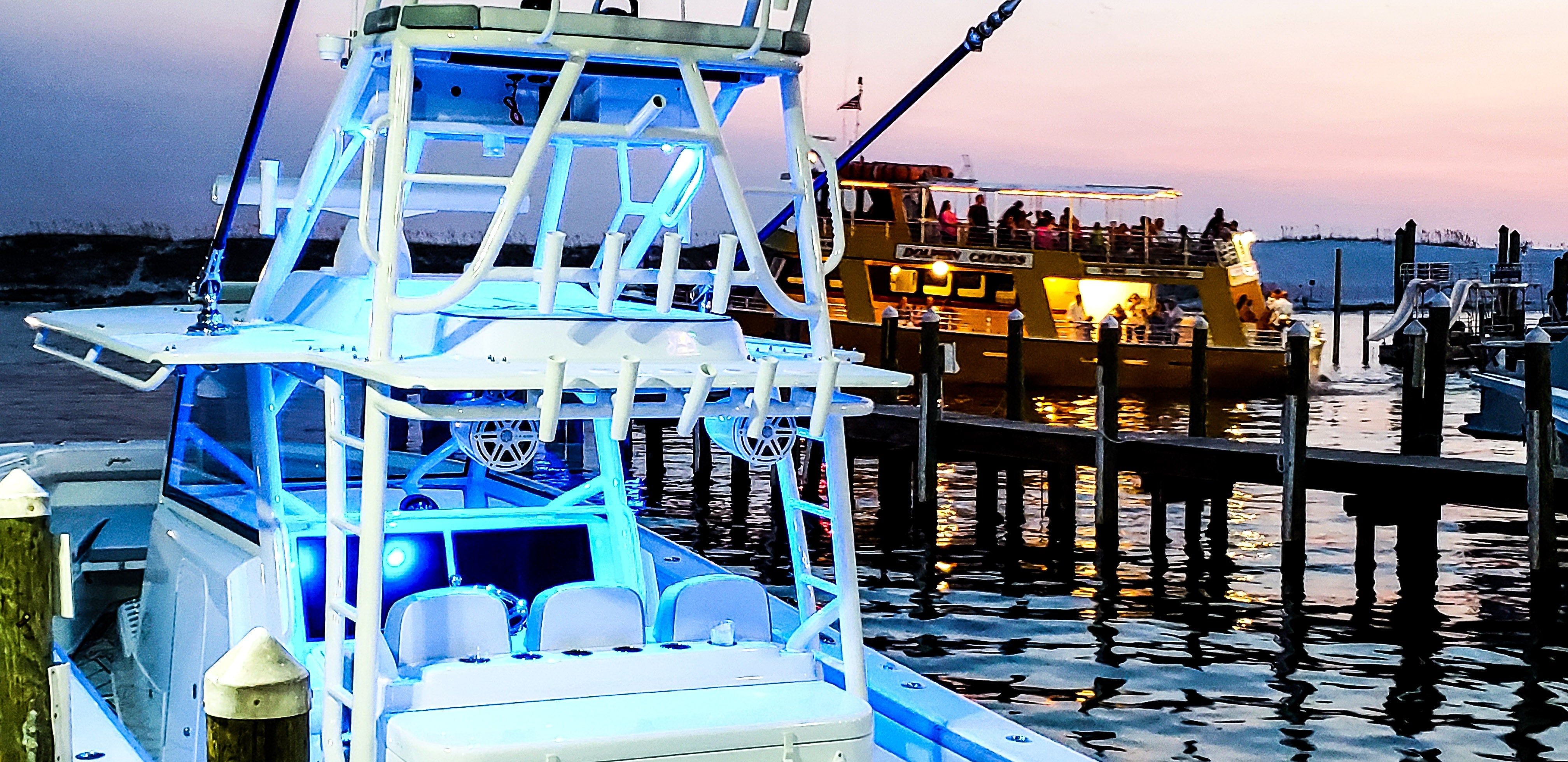 Sportfishing boat with blue underglow and rod holders docked at a pier, with a Dolphin Cruises passenger boat at dusk.