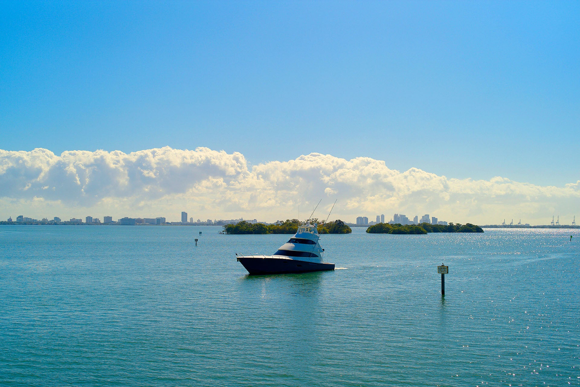Sportfishing yacht cruising across a calm bay with a city skyline and mangrove islands on the horizon.